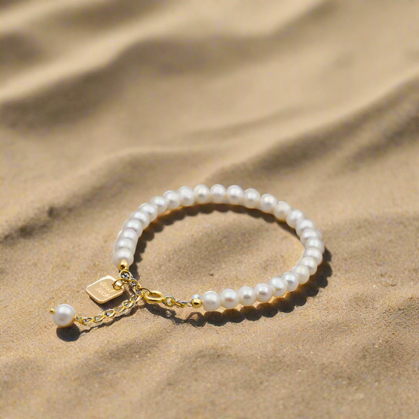 Round pearl titanium steel beaded bracelet on sand with gold chain and charm.